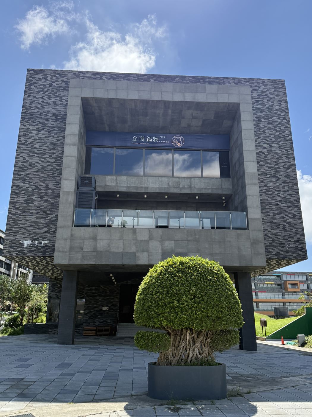 KIRI 國際原住民族文創園區外觀，深色石材方塊量體，二樓掛著金時鍋物招牌，門口有一棵球狀造型老榕樹